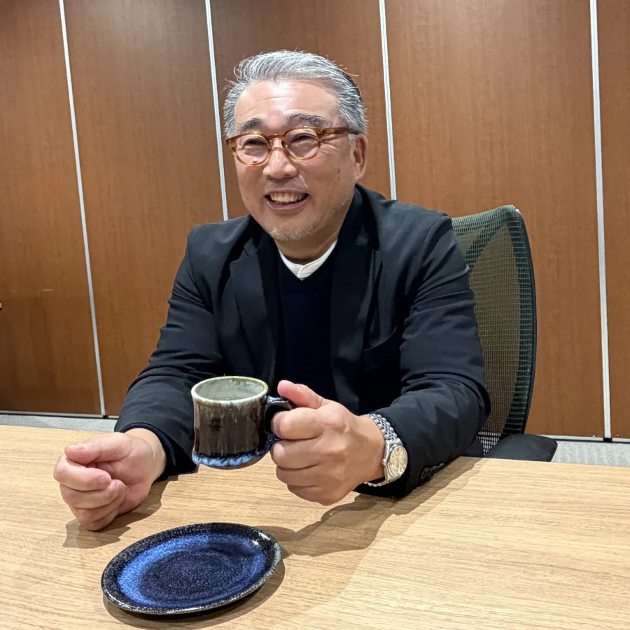 片野秀樹氏が温かい飲み物を片手に微笑む様子